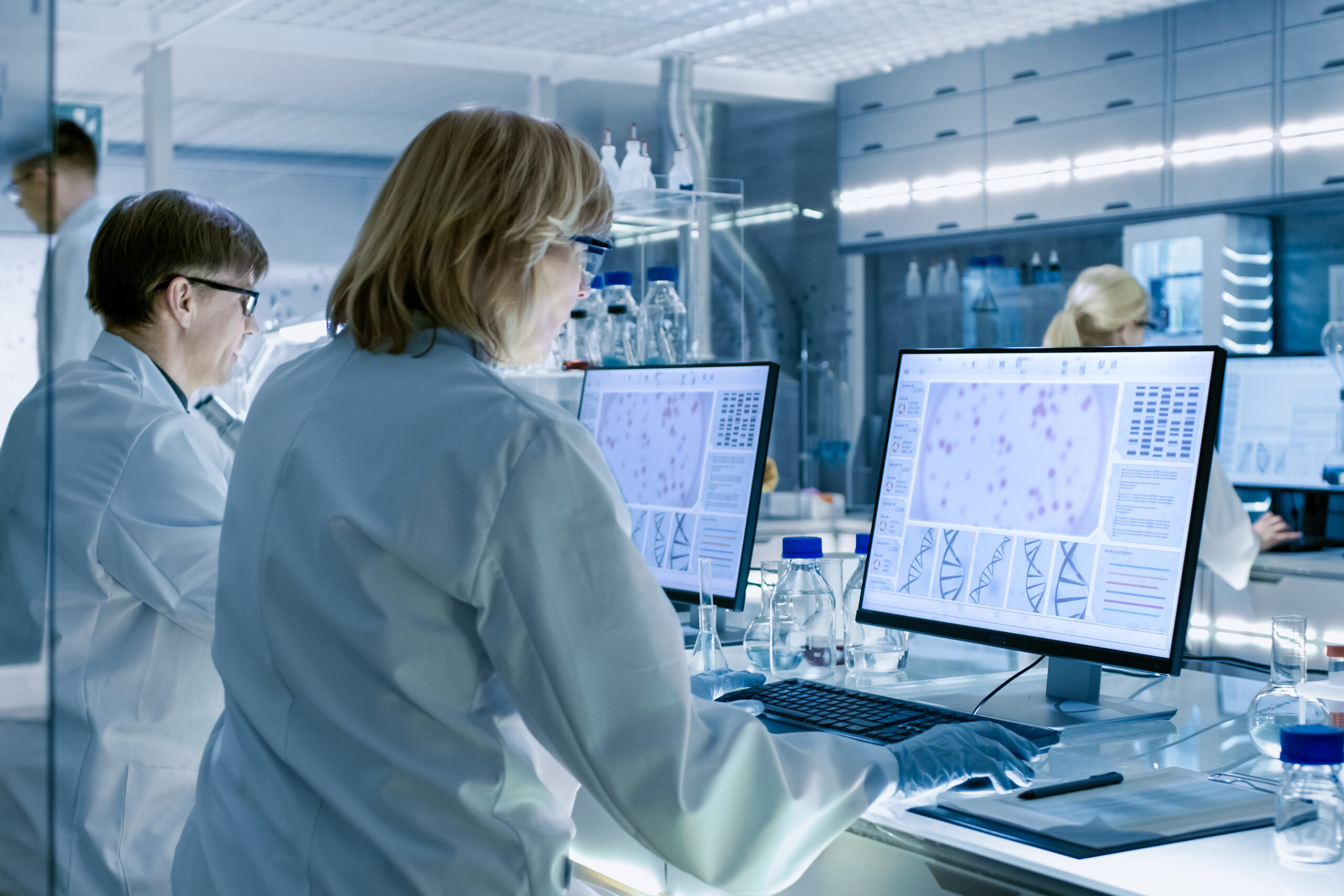 Female and Male Scientists Working on their Computers In Big Modern Laboratory. Various Shelves with Beakers, Chemicals and Different Technical Equipment is Visible.